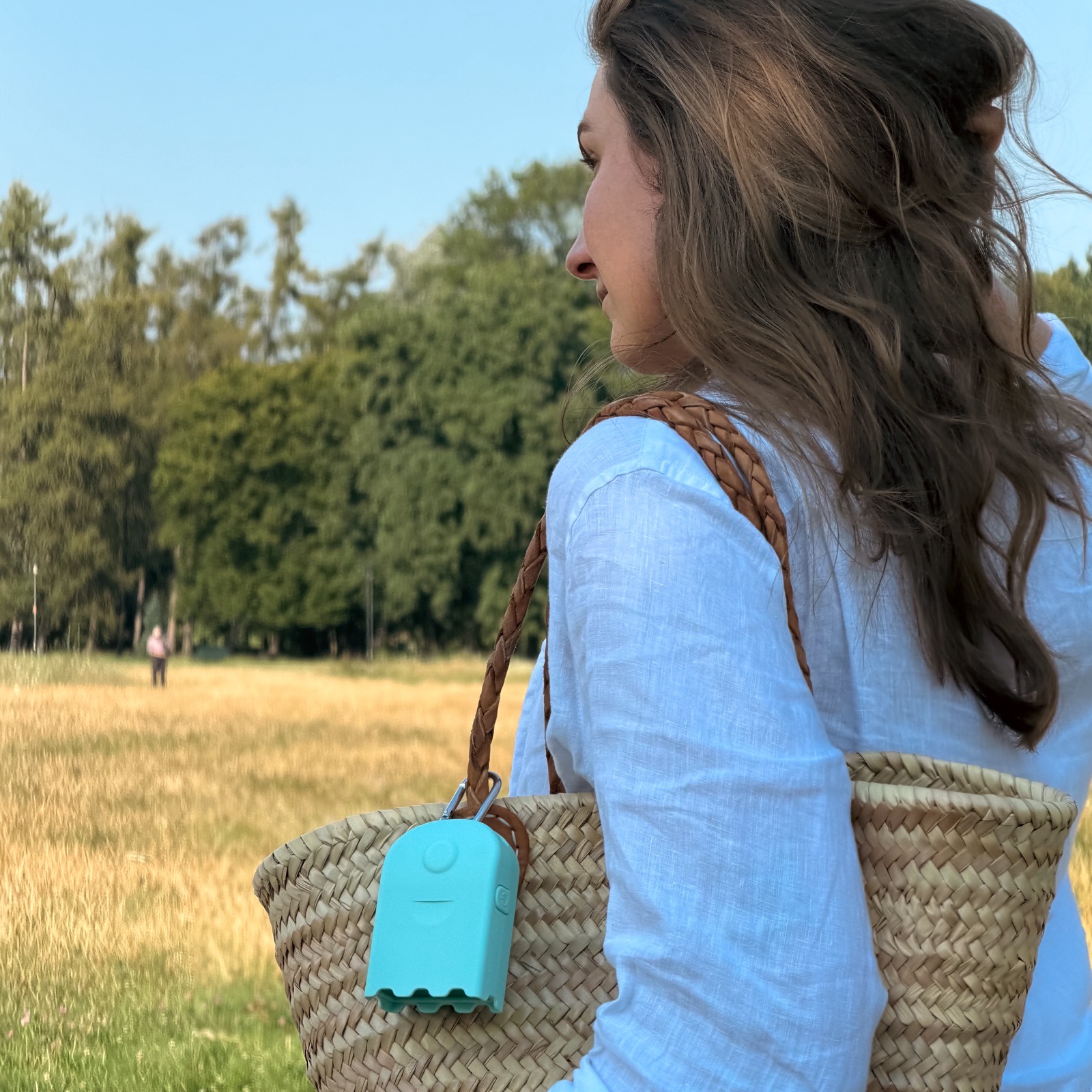 A woman carrying a straw tote bag with an AirBeam Mini clipped to it, walking through a sunny field